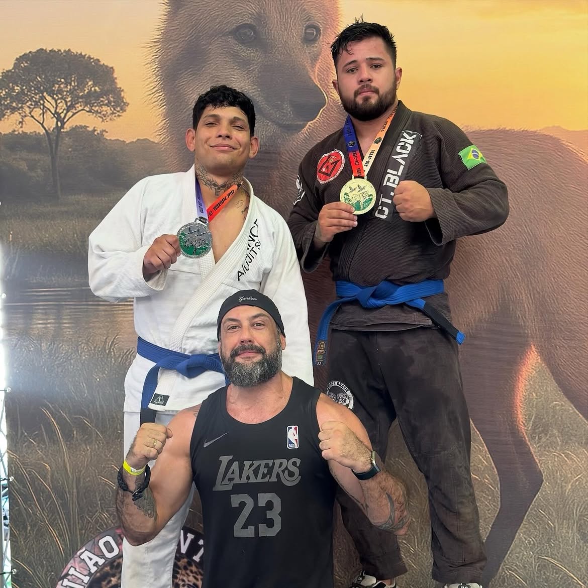 Dois atletas de kimono exibindo medalhas de ouro e prata atrás do professor, que ergue os punhos