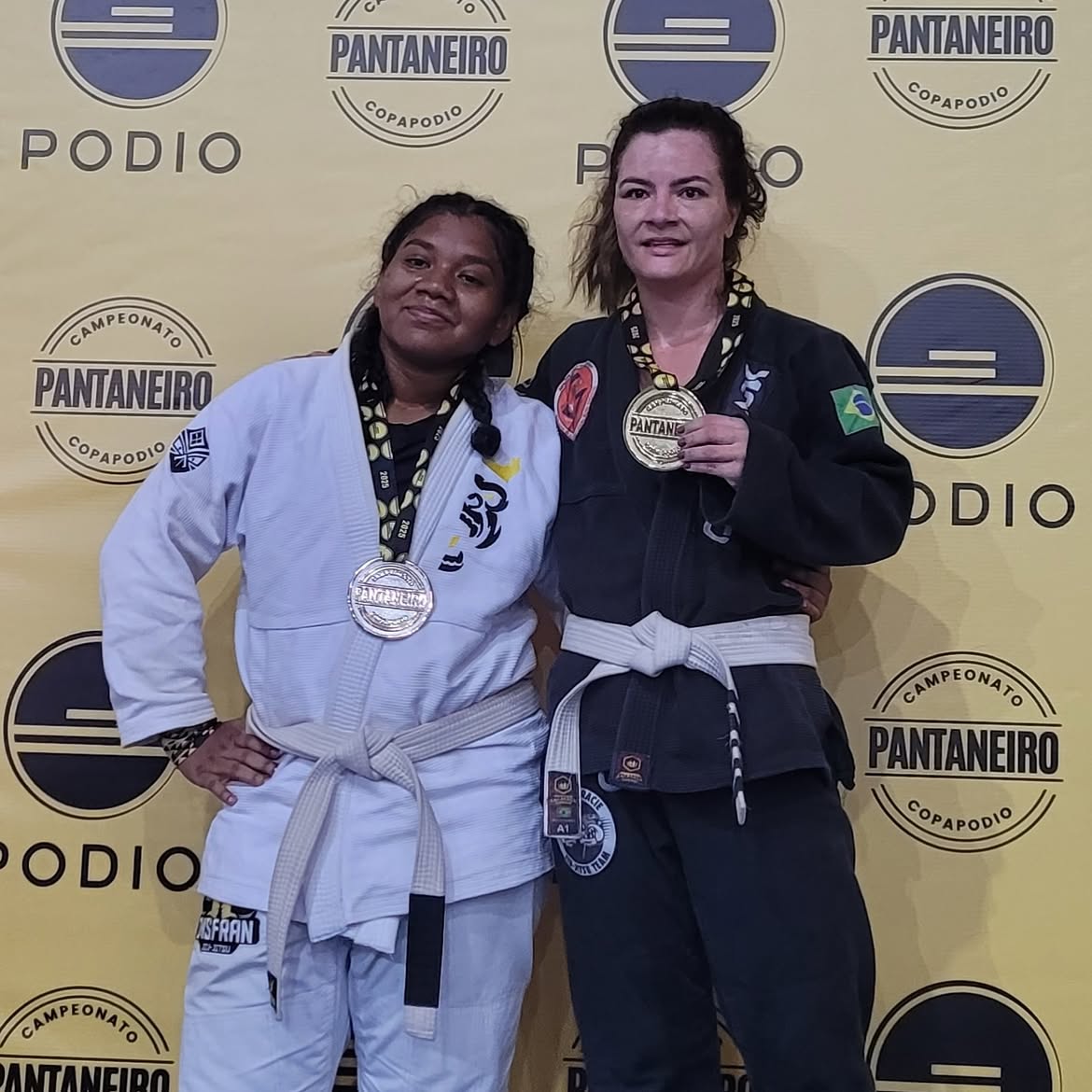Duas atletas de kimono lado a lado exibindo medalhas de ouro e prata no painel do campeonato