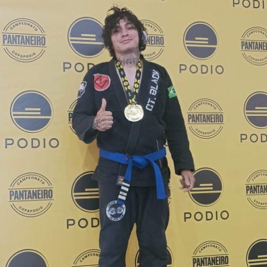 Atleta de kimono preto e faixa-azul com medalha de ouro, dando joinha em frente ao painel do campeonato