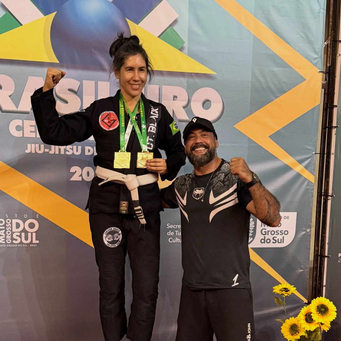 Atleta no pódio com duas medalhas de ouro e o punho erguido, ao lado do professor
