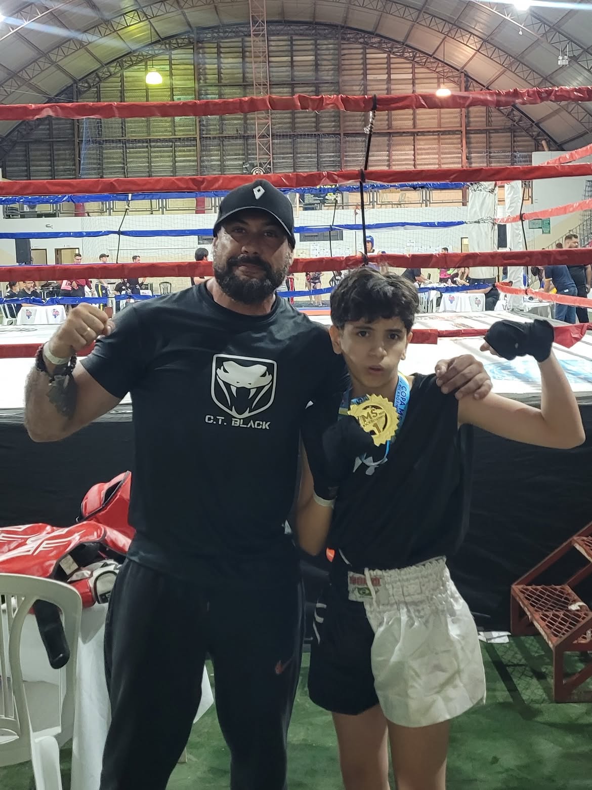 Professor e jovem atleta posando com medalha junto ao ringue