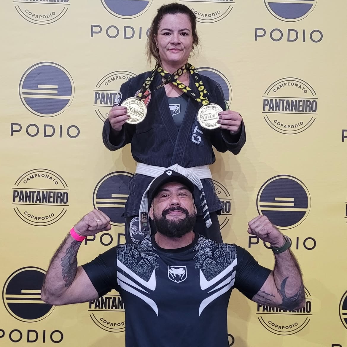 Atleta de kimono preto com duas medalhas de ouro ao lado do professor, em frente ao painel do campeonato