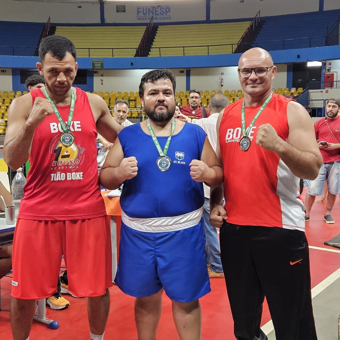 Três atletas com medalhas e punhos erguidos dentro do ginásio de competição